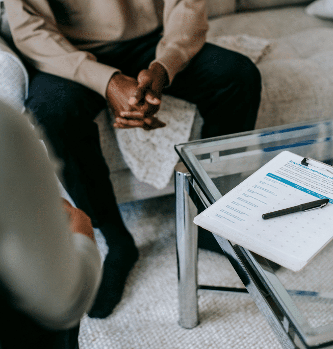 Two people sit across from each other in a living room. One person’s hands are clasped, while a clipboard with a form and pen rests on a glass table between them, suggesting a counseling or therapy session.