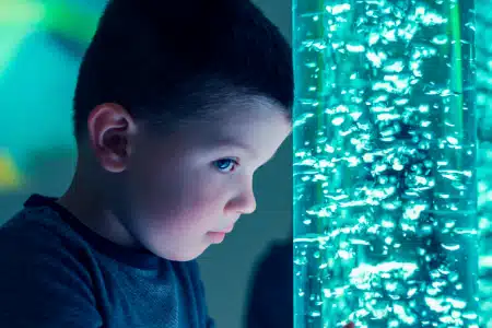 A young boy with short dark hair closely observes a tall, illuminated bubble tube filled with rising bubbles, his face lit by the blue glow in a dimly lit room.